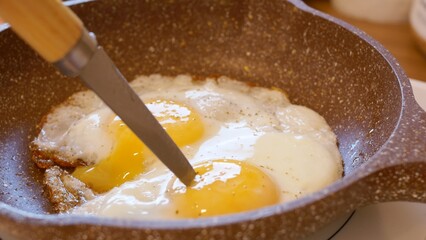 Close-up of frying pan with scrambled eggs, the yolk is pierced with knife for deep frying. Egg breakfast is quick and nutritious. Habitual preparation of scrambled eggs for breakfast is daily routine