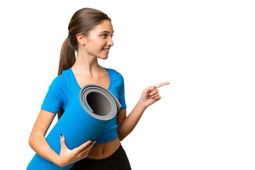 Teenager caucasian girl going to yoga classes while holding a mat over isolated background pointing to the side to present a product © luismolinero
