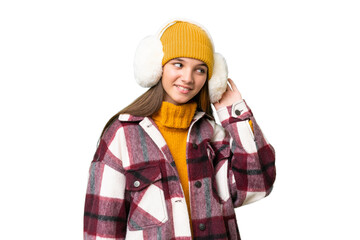 Teenager caucasian girl wearing winter muffs over isolated background listening to something by putting hand on the ear