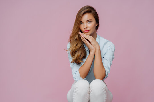 Serious beautiful woman with long hair, dressed in shirt and trousers, keeps palms pressed together undr chin, has confident calm look, sits on chair over violet background, copy space area aside