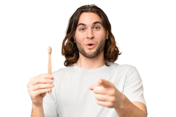 Young handsome man brushing teeth over isolated background surprised and pointing front