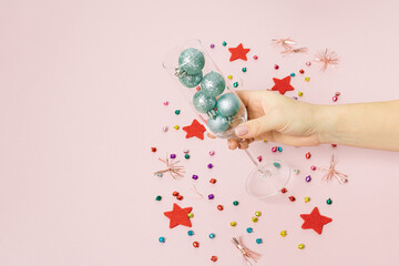 Champagne glass with baubles in hand decorated with colorful confetti and red star on pastel pink background. Christmas or New Year party concept. Minimal winter holiday season idea with copy space.