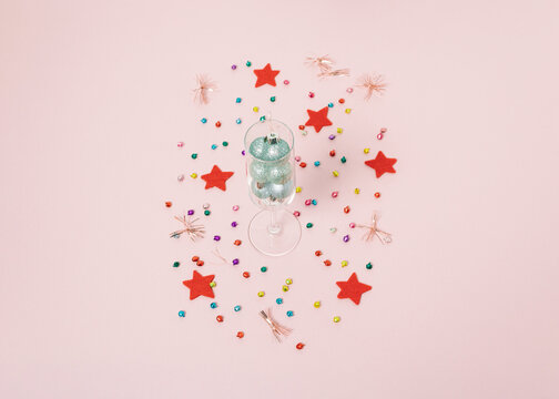 Champagne glass with baubles decorated with colorful confetti and red star on bright pastel pink background. Christmas or New Year party concept. Minimal winter holiday season idea.