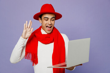 Young smiling fun middle eastern IT man 20s he wear white turtleneck red hat scarf hold use work on laptop pc computer waving hand get video call talk isolated on plain pastel light purple background.