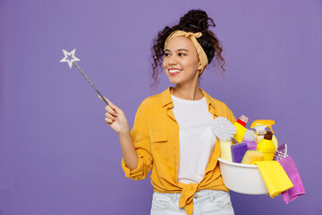 Young housekeeper woman in yellow shirt tidy up hold basin with detergent bottles point magic wand...