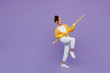 Fototapeta na wymiar Full body side view fun young housekeeper woman wear yellow shirt tidy up hold broom sweeping brush look aside clean house isolated on plain pastel light purple background studio. Housework concept.