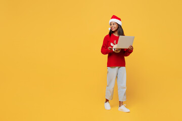 Merry IT little kid teen girl 13-14 year old wear red xmas sweater with deer Santa hat posing work hold use laptop pc computer isolated on plain yellow background. Happy New Year 2023 holiday concept