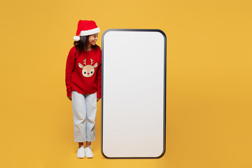 Merry fun little kid teen girl 13-14 years old wear red xmas sweater Santa hat posing look at big blank screen mobile cell phone with area isolated on plain yellow background. Happy New Year concept.