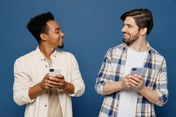 Young two friends cheerful smiling happy cool men 20s wear white casual shirts together hold in hand use mobile cell phone chatting look to each other isolated plain dark royal navy blue background.