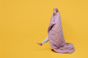 Full body side view young fun woman wrapped in blanket duvet wears purple pyjamas jam sleep eye...