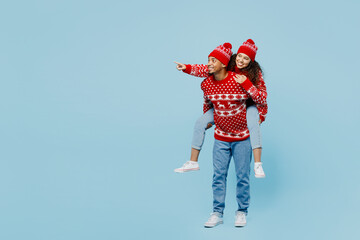 Full body merry young couple two man woman wear red Christmas sweater Santa hat posing give piggyback ride to joyful sit on back point aside isolated on plain blue background. New Year 2023 concept.