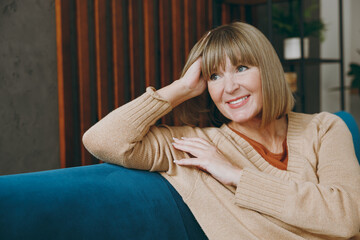 Elderly fun minded dreamful happy woman 50s years old wears casual clothes sits on blue sofa look aside prop up head stay at home flat rest relax spend free spare time in living room indoor grey wall.