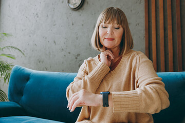 Elderly woman 50s year old wear casual clothes sit on blue sofa use look at smart watch put hand on...