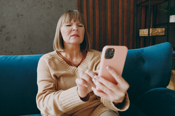 Elderly woman 50s years old wears casual clothes sits on blue sofa hold use mobile cell phone without glasses squint stay at home flat rest relax spend free spare time in living room indoor grey wall.