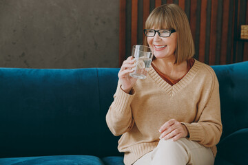 Elderly smiling happy fun woman 50s years old wears casual clothes glasses sits on blue sofa hold glass drink water stay at home flat rest relax spend free spare time in living room indoor grey wall. - Powered by Adobe