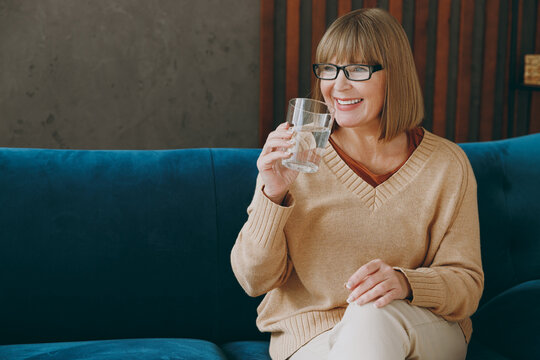 Elderly smiling happy fun woman 50s years old wears casual clothes glasses sits on blue sofa hold glass drink water stay at home flat rest relax spend free spare time in living room indoor grey wall.