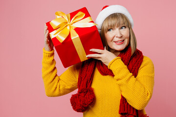 Merry elderly woman 50s years old wear knitted sweater red scarf Santa hat posing hold present box ribbon bowgift look aside isolated on plain pink background. Happy New Year Christmas 2023 concept.