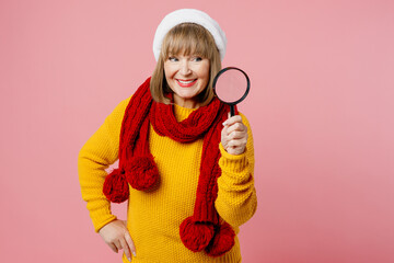 Merry fun elderly woman 50s years old wear yellow knitted sweater red scarf Santa hat posing hold use reading-glass magnifier isolated on plain pink background. Happy New Year Christmas 2023 concept.