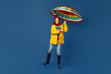 Full body young woman in sweater red hat yellow waterproof raincoat outerwear hold opened umbrella jump high isolated on plain dark royal navy blue background Outdoor wet fall weather season concept