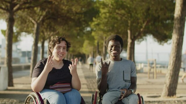 Happy ouple using wheelchairs showing positive gestures. Portrait of Caucasian woman and African American man looking at camera and smiling in park. Couple with disability concept