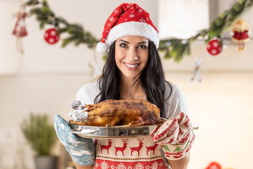 Satisfied young woman in a Christmas apron and Santa hat holding a roasting pan with a roast goose...