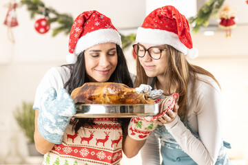 Two cheerful cooks, mom and her teenage daughter, in Christmas aprons with Santa hats, have baked a goose or turkey, they are smelling it