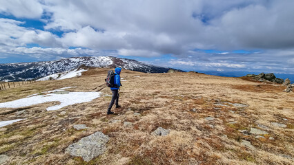 Rear view of man with hiking backpack walking on alpine meadow from Ladinger Spitz to Gertrusk, Saualpe, Lavanttal Alps, Carinthia, Austria, Europe. Trekking on cloudy early spring day. Wanderlust