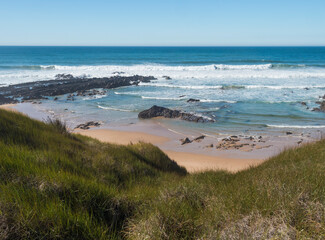 View of empty Praia da Angra das Melancias beach with ocean waves, stone, wet golden sand and green vegetation at wild Rota Vicentina coast near Vila Nova de Milfontes, Portugal.