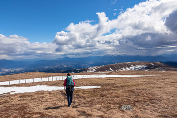 Rear view of woman with hiking backpack on alpine meadow walking along fence from Ladinger Spitz to Gertrusk, Saualpe, Lavanttal Alps, Carinthia, Austria, Europe. Trekking on cloudy early spring day