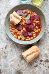 Plate of chorizo and bean stew with crusty bread on a light-brown granite background, vertical shot, above view