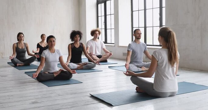 Rear view female yogini coach and multi-ethnic girls and guys practising yoga session seated in lotus pose meditating indoors, feeling inner calmness, increasing body energy flow, stress and anxiety