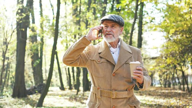 Slow-motion mature grey-haired successful retired man walking in autumn park talking on phone holding cup of coffee wearing beige coat. Elegant senior grandfather using smartphone communicating.