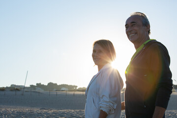 Fototapeta na wymiar Happy mature couple standing on beach on summer day and smiling. Medium shot of cheerful senior man and woman in sportswear looking forward, spending time together. Sport, family concept