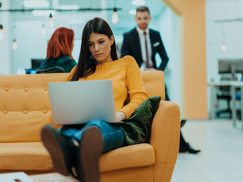 Handsome businesswoman (employee) sitting on a yellow couch while working on her laptop in startup company.