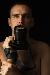 Close-up of a man with a camera against a black background. Photographer holding camera at mouth level. Breast portrait of a Caucasian man with a DSLR camera shirtless. Low key.