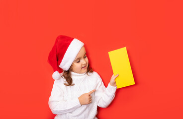 Cute caucasian kid girl in red Santa's hat points with his finger on certificate or greeting card empty copy space for design or text. Banner. Christmas mockup template.