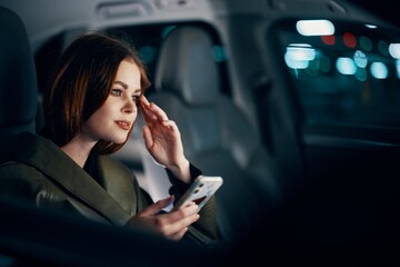  stylish, luxurious woman in a leather coat sitting in a black car at night on the passenger seat, looking thoughtfully to the side, holding her hand near her face, and the other holding a smartphone