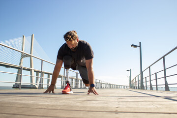 Portrait of man with leg prothesis on start for running. Dark-haired man standing on bridge over sea bending touching ground with hands looking down. Health and life of people with disability concept