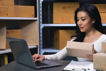 A female employee is packing parcels and checking customer addresses with a laptop to deliver...
