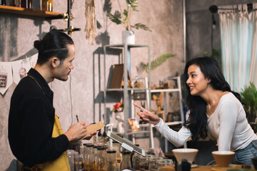 Barista discussing coffee and food menus with customers, Asian smiling woman client standing in front of counter for ordering coffee to takeaway orders in the morning at a cafe shop.