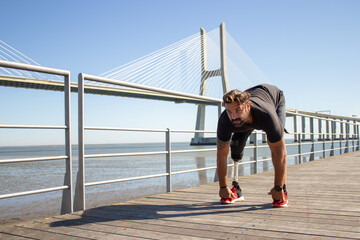 Portrait of man with disability running in morning. Serious man in casual clothes standing on bridge over sea bending his body getting ready to run quick. Active life of people with disability concept