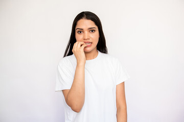 Portrait of nervous young woman biting nails in anxiety over white background. Caucasian lady wearing white T-shirt feeling worried and uneasy. Stress or fear concept