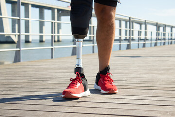Close-up of legs of man with disability walking along bridge. Sporty man in casual clothes walking away after training looking at blue sea. Health, active lifestyle of people with disability concept
