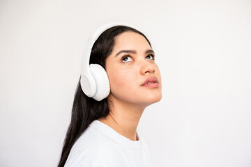 Close-up of serious young woman in headphones. Portrait of Caucasian lady wearing white T-shirt listening to music and looking up over white background. Music and leisure concept