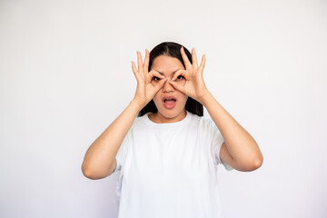 Portrait of surprised young woman making glasses with fingers. Caucasian lady wearing white T-shirt peeking through hands over white background. Astonishment and awe concept