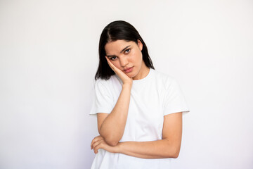 Portrait of bored young woman leaning head on hand over white background. Caucasian lady wearing white T-shirt looking at camera with indifference. Boredom concept