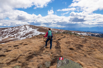 Woman walking next to path mark with Austrian flag painted on rock on remote alpine meadow near Ladinger Spitz, Saualpe, Carinthia, Austria, Europe. Hiking trail in the Austrian Alps in early spring