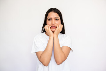 Portrait of neurotic young woman biting nails over white background. Caucasian lady wearing white T-shirt looking at camera in anxiety or fear. Stress and depression concept