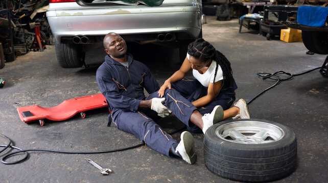 African American woman mechanic helped a black engineer man with knee pain due to work lie on the garage floor. - Powered by Adobe