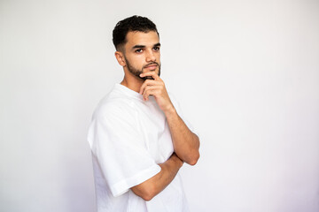 Portrait of focused young man thinking. Pensive Caucasian male model with dark hair and beard in white T-shirt looking away with hand on chin. Focus, contemplation concept